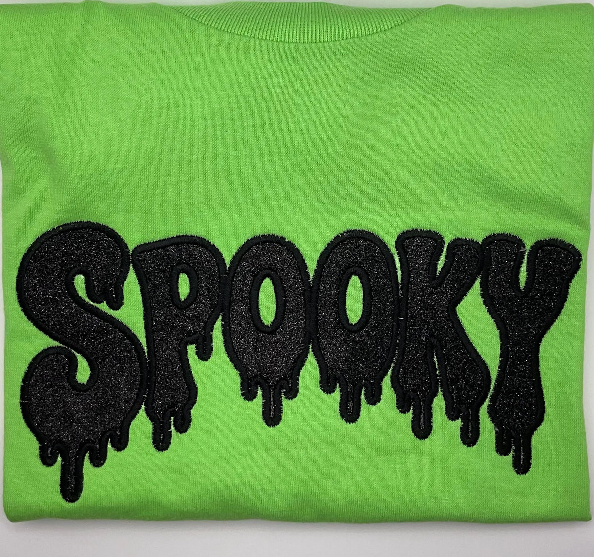 A folded lime green sweatshirt featuring a large, black glittery dripping 'SPOOKY' text graphic across the chest.