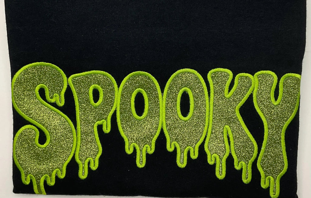 A close-up of a black fabric item featuring the word 'SPOOKY' in large, glittery lime green letters with a dripping outline.
