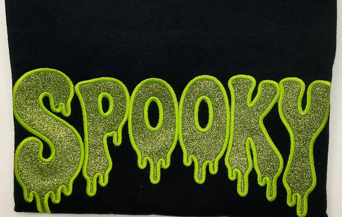 A close-up of a black fabric item featuring the word 'SPOOKY' in large, glittery lime green letters with a dripping outline.