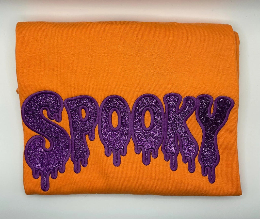 An tangerine folded sweatshirt featuring a large purple glittery 'SPOOKY' text design with a dripping effect, displayed on a plain white background.